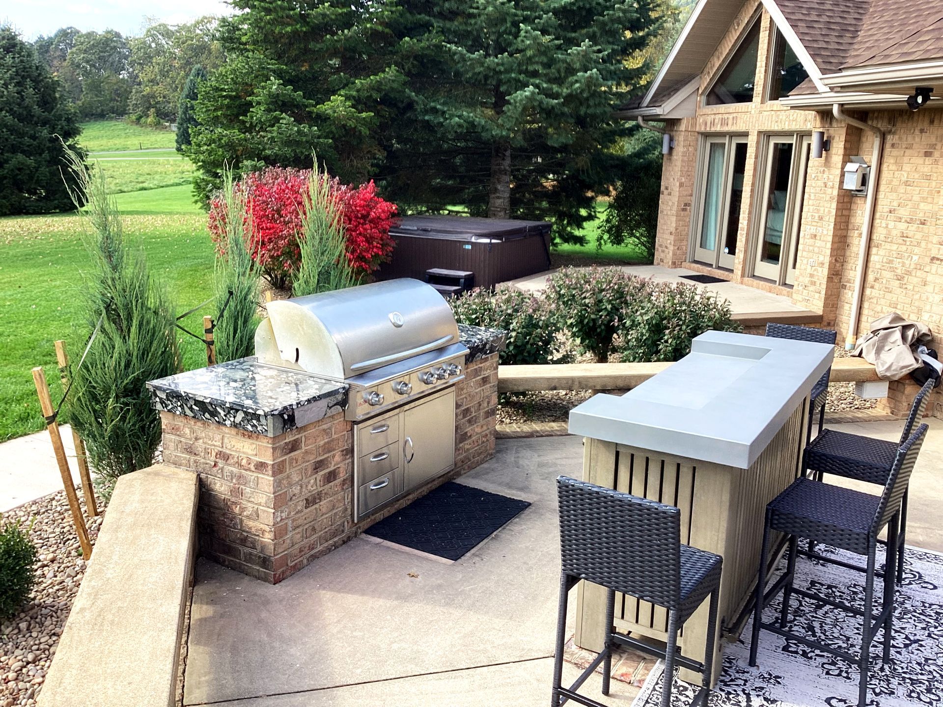 Outdoor kitchen with grill, bar seating, hot tub, and landscaping near a house.
