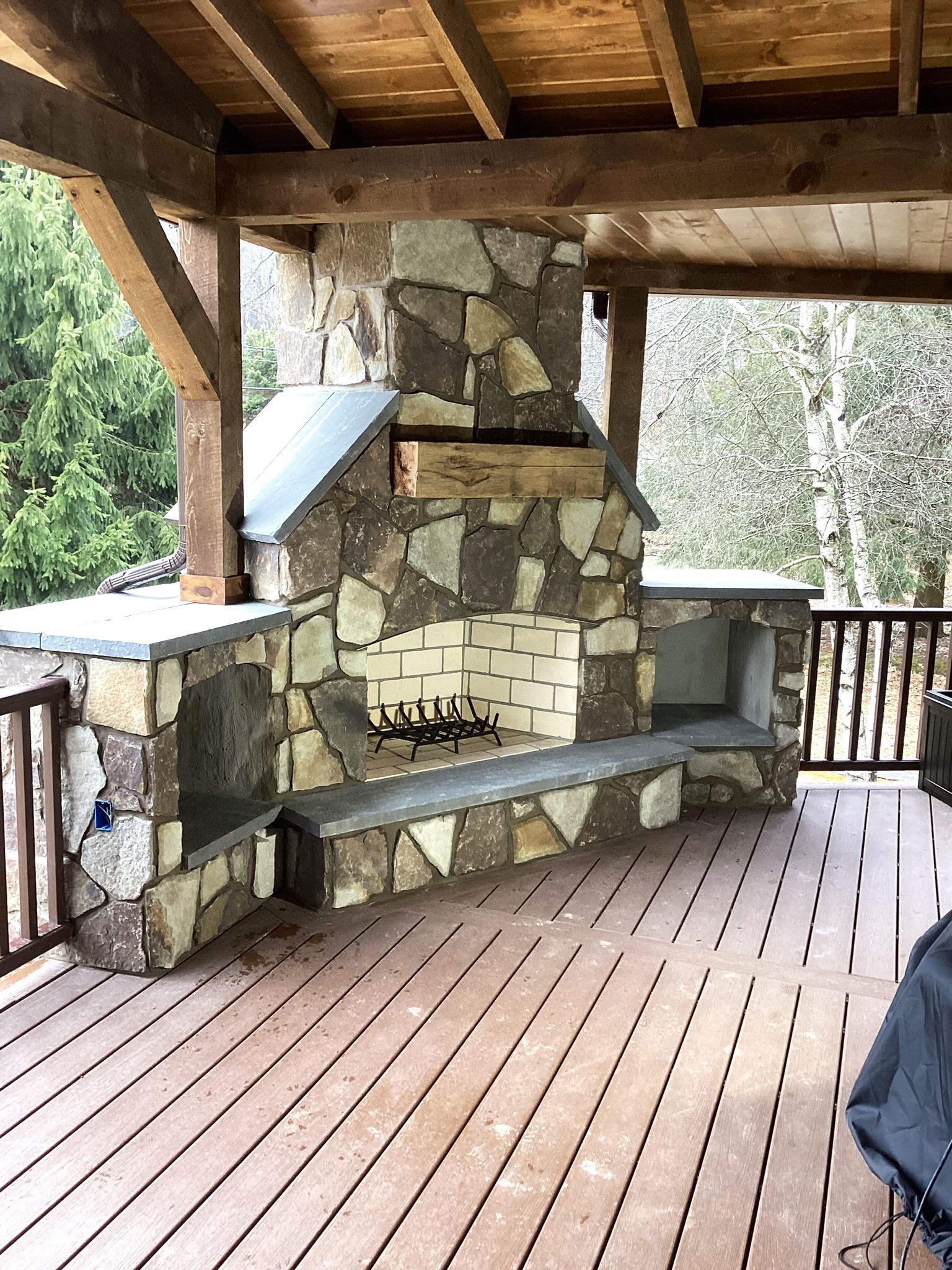 Stone outdoor fireplace on a wooden deck under a wood-beamed roof.