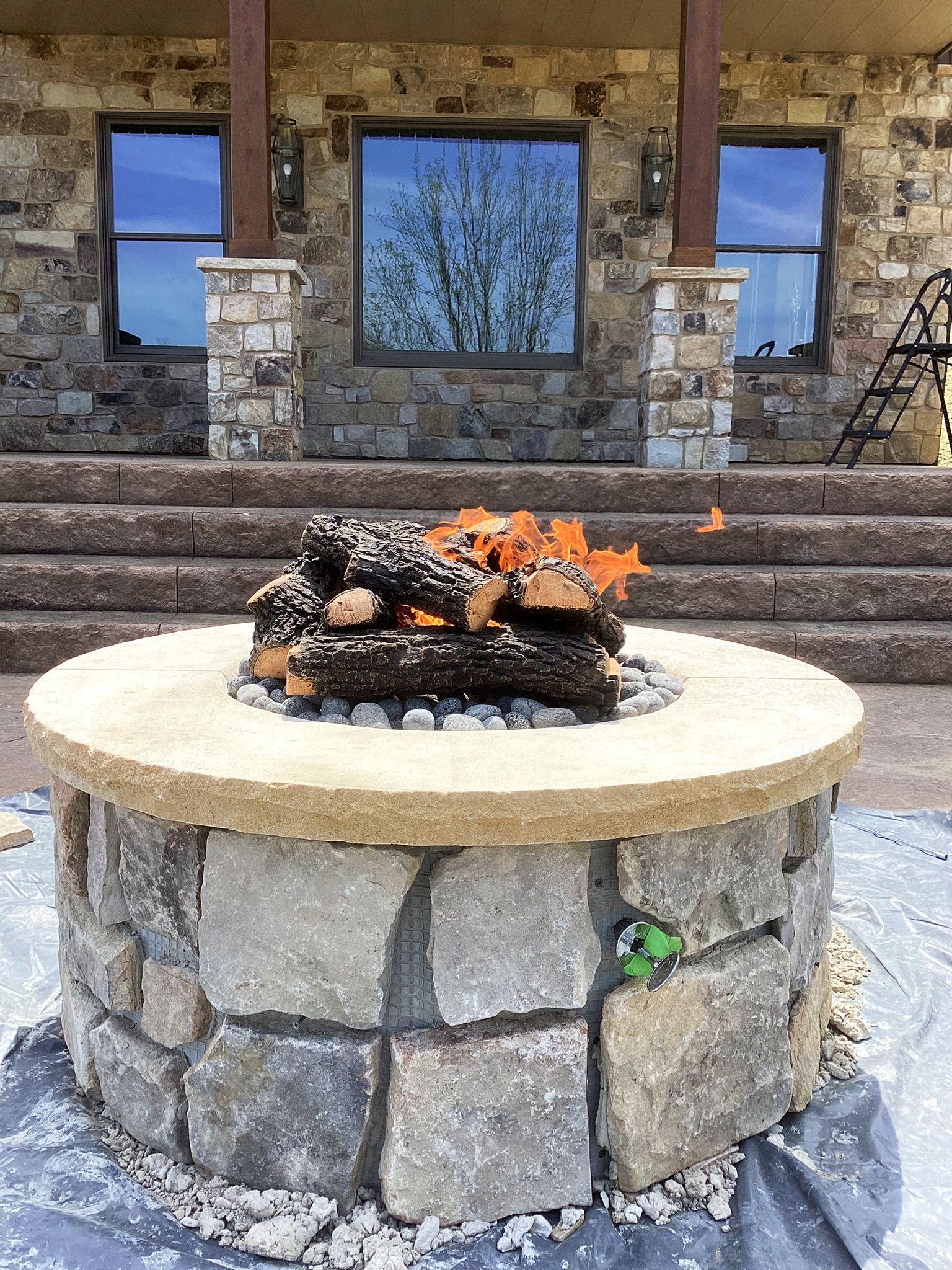 Fire pit with burning logs, built with stone and concrete, set in front of a house with stone exterior.