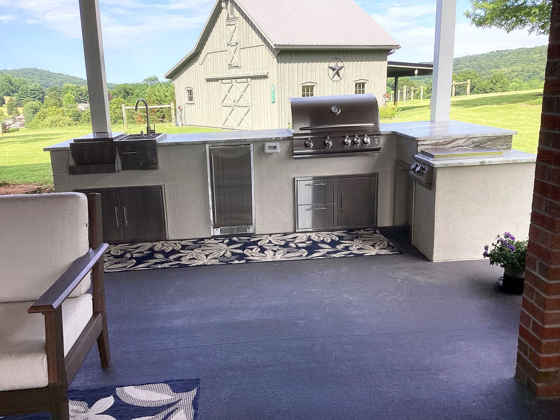 Outdoor kitchen with stainless steel appliances, a barn in the background, and a seating area on a patio.