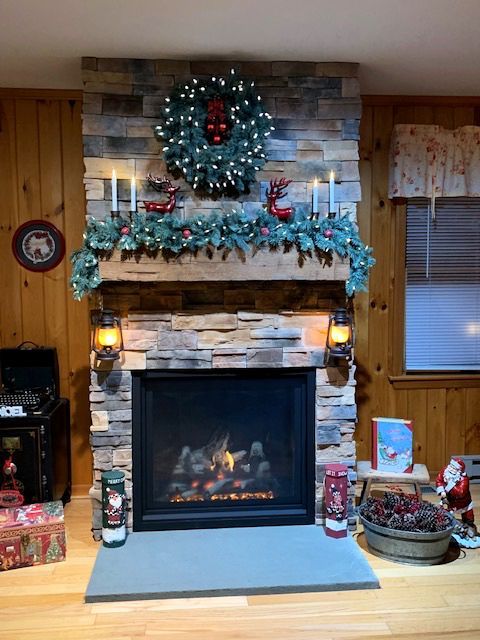 Christmas-decorated stone fireplace with wreath, garland, candles, and lanterns in a cozy, wooden-paneled room.