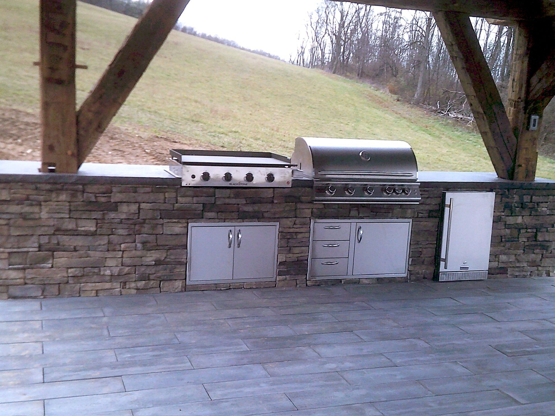 Outdoor kitchen with stainless steel grill and appliances set in stone wall, beneath a wooden pergola.