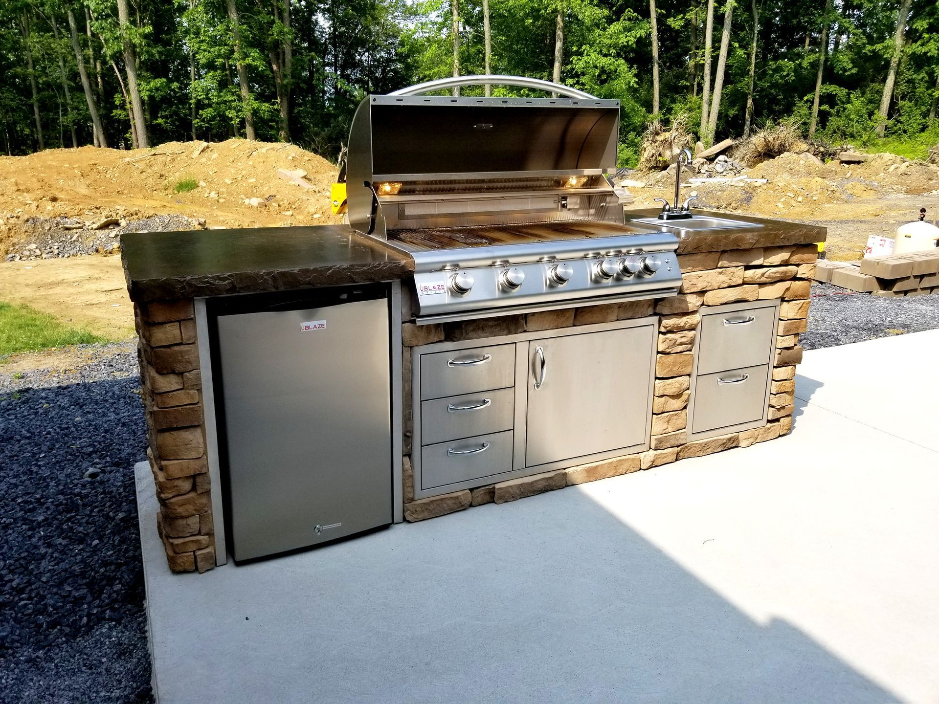 Outdoor kitchen with stainless steel grill and appliances, stone facade, on a concrete patio.