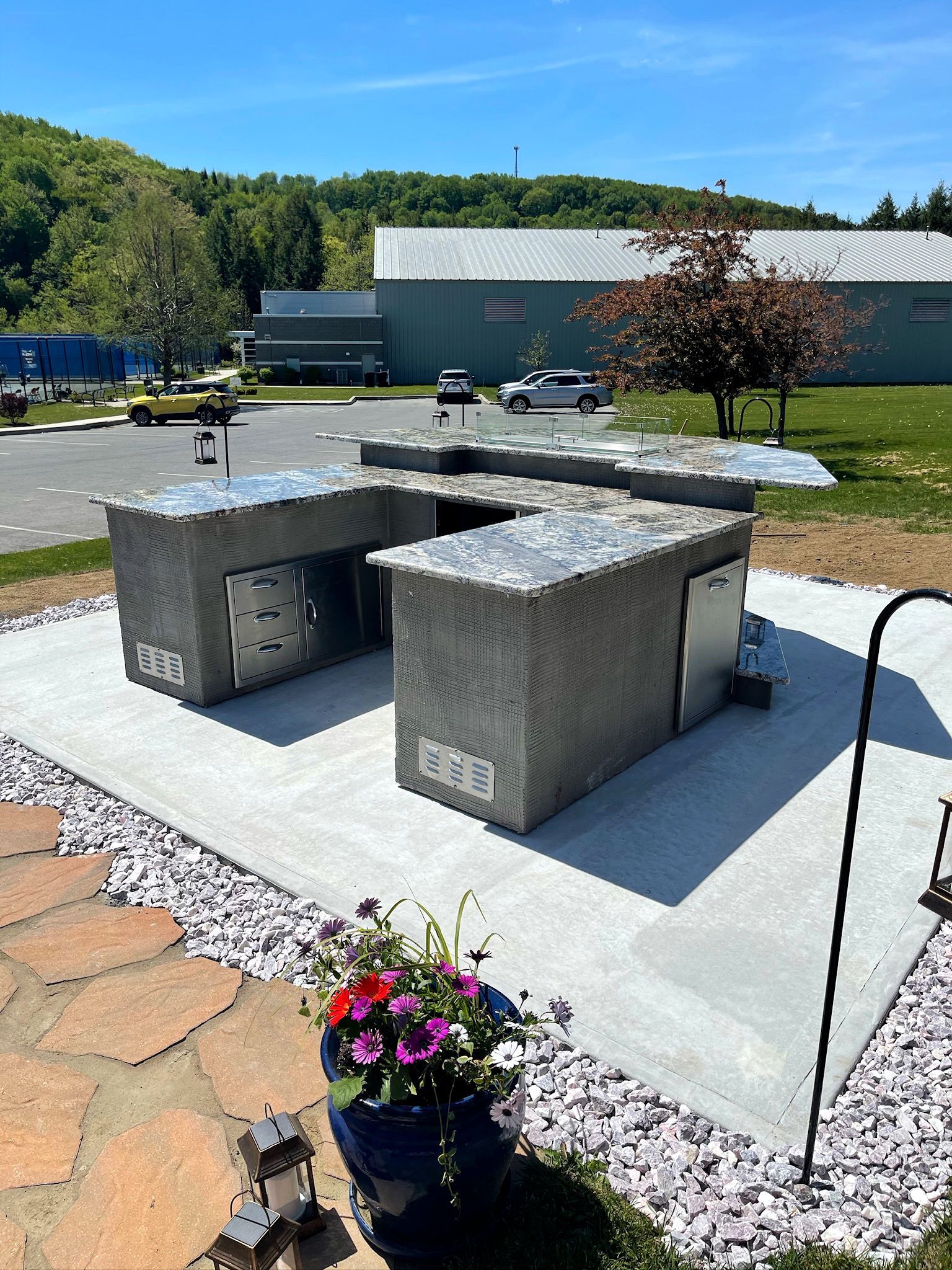 Outdoor concrete and metal structure, possibly a grill or kitchen, with flowers in the foreground and buildings in the background.