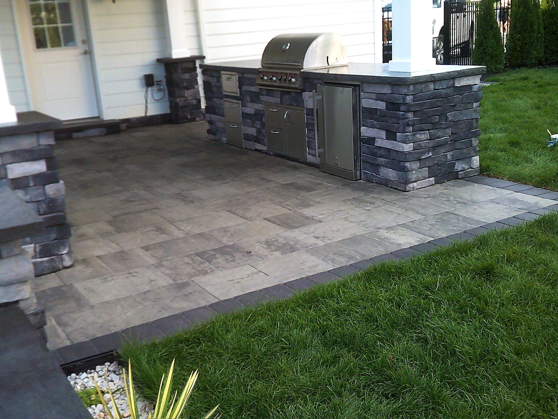Outdoor kitchen with grill, refrigerator, and stone facade on a paved patio, next to green grass.
