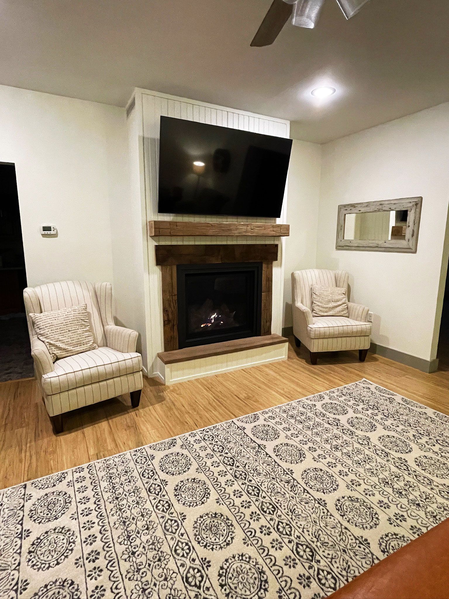 Living room with fireplace, TV, two armchairs, rug, and mirror. Warm colors and wooden accents.