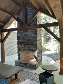 Outdoor stone fireplace under a wooden patio roof. A rug, table, and seating are nearby.