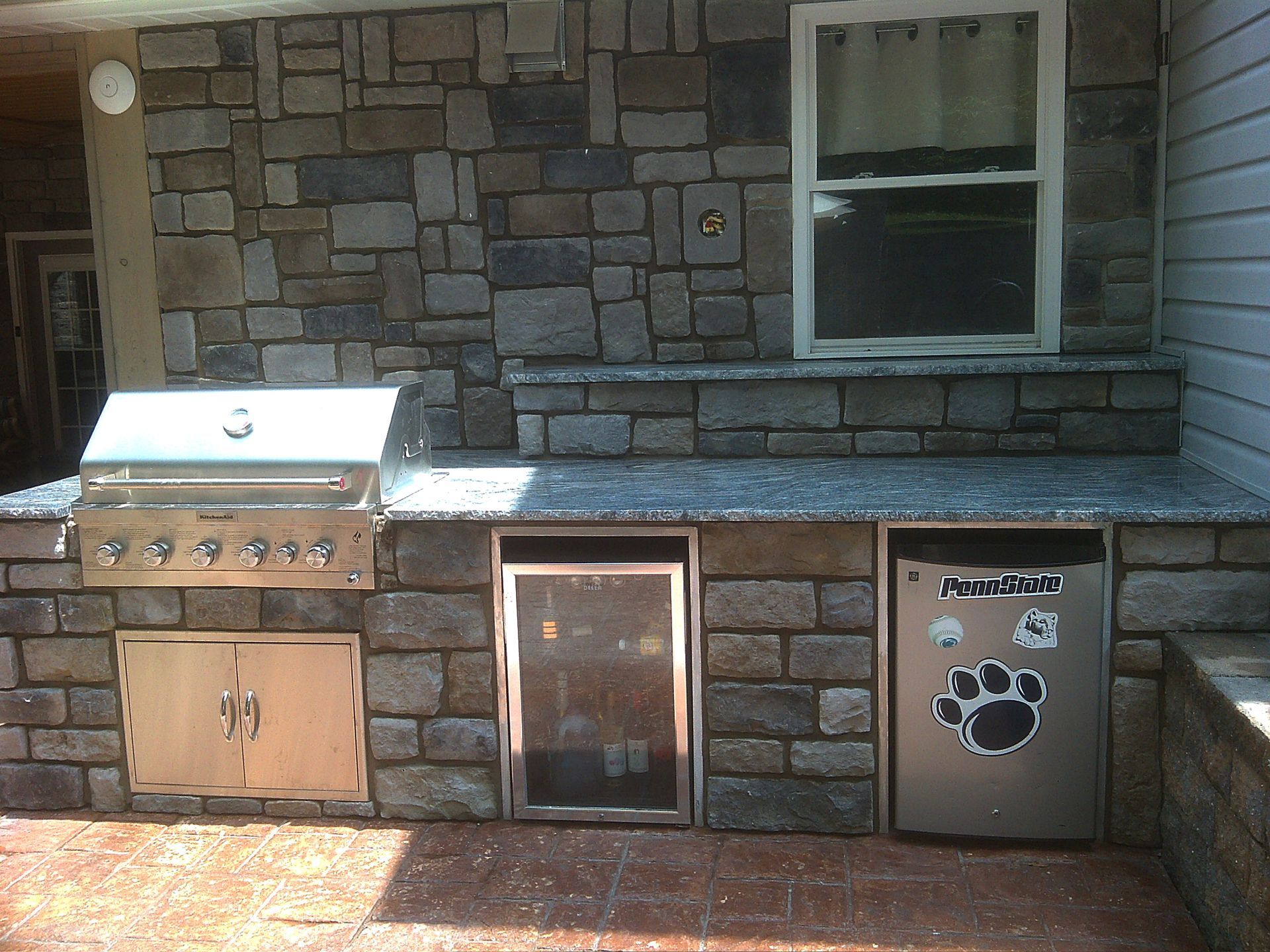 Outdoor kitchen with stone veneer, stainless steel grill, refrigerator, and cabinets.