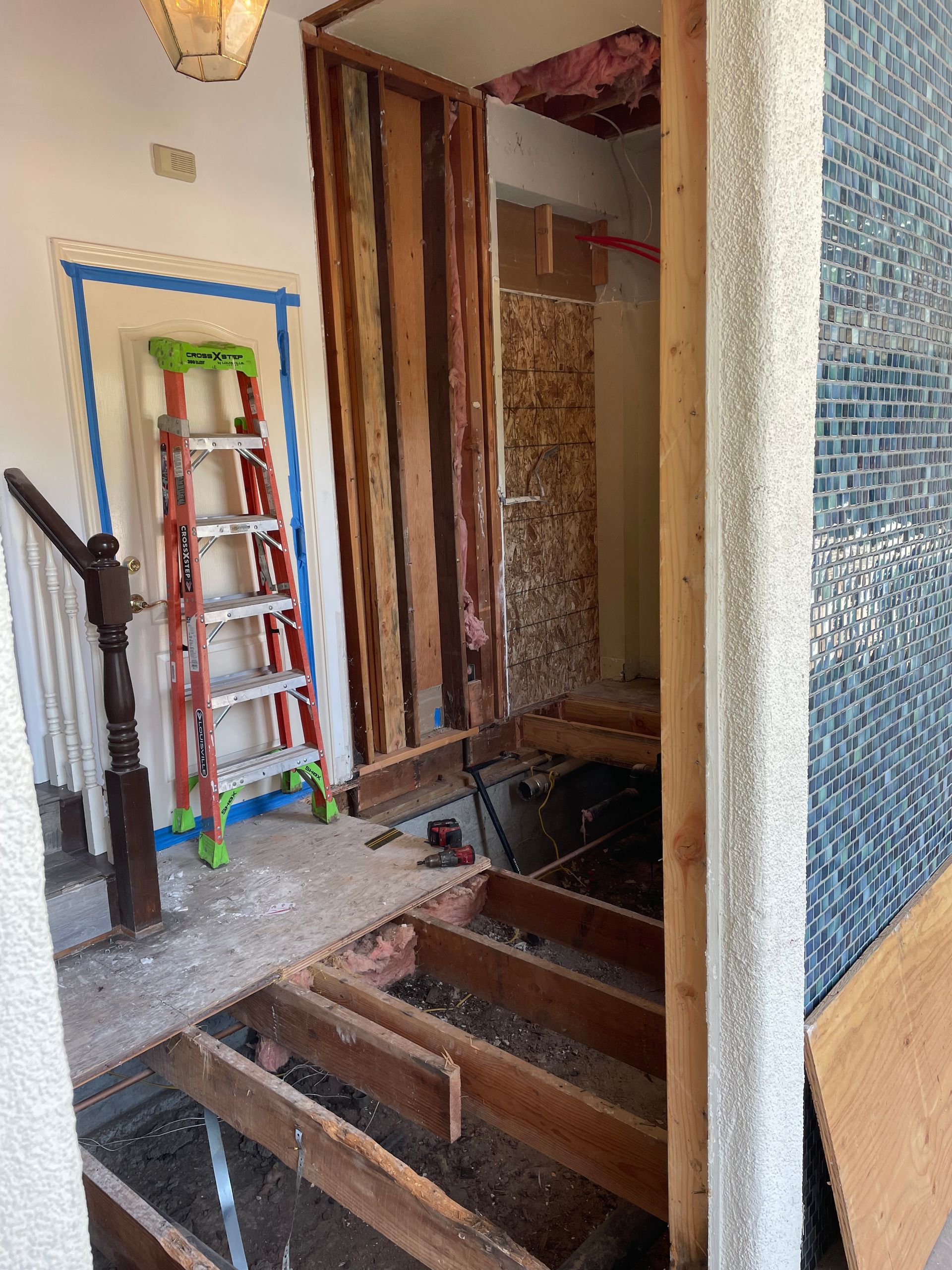 Staircase under renovation with exposed floor joists, a ladder, and a doorway framed by wood and tile.