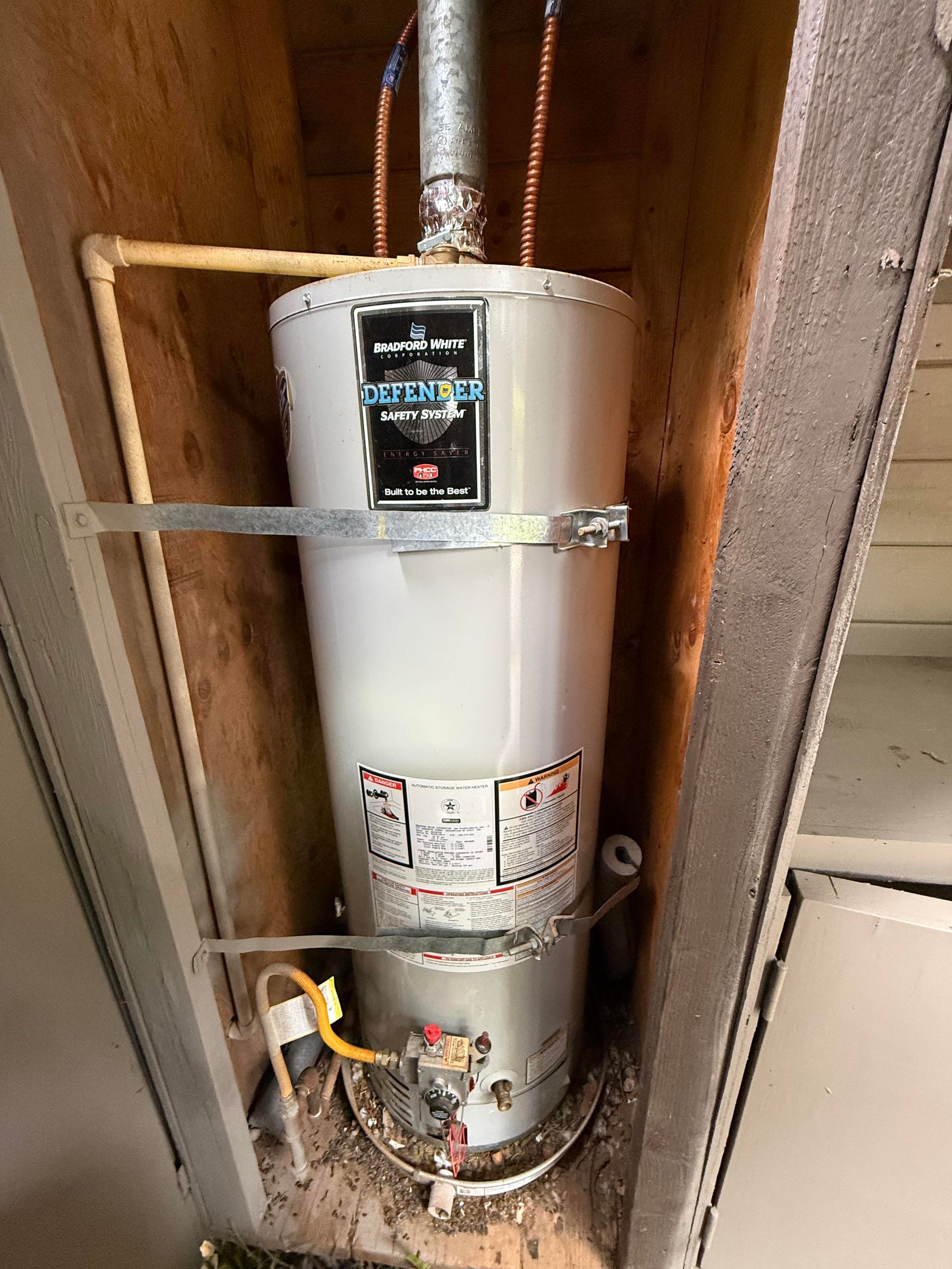 White water heater in a wooden closet with exposed pipes and metal straps, front labels visible