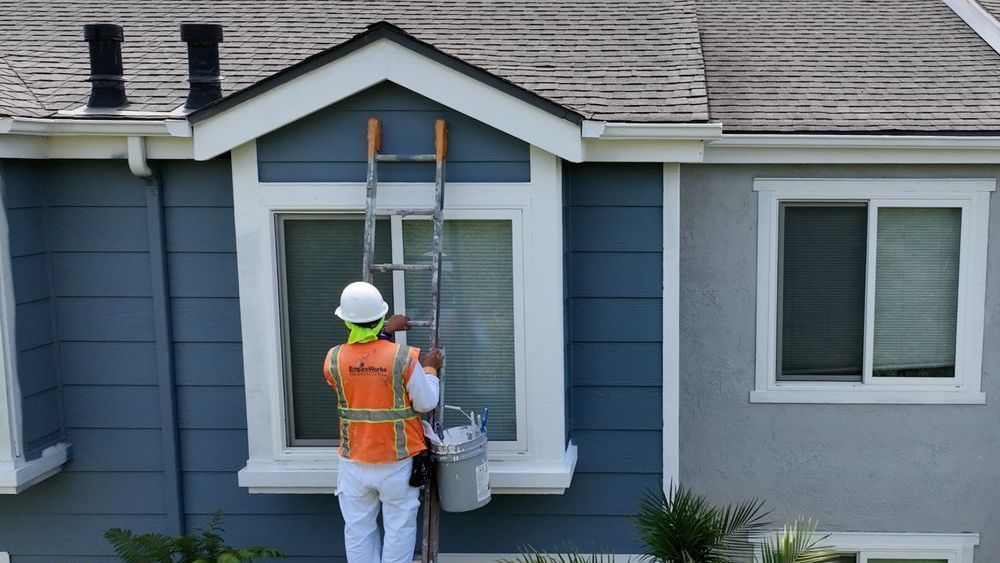 Person on ladder painting window frame of blue house.
