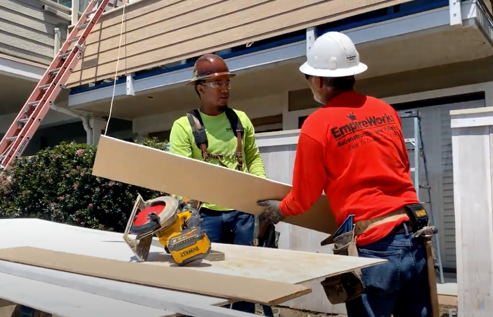 Two construction workers installing siding, one holding a board, near a circular saw.