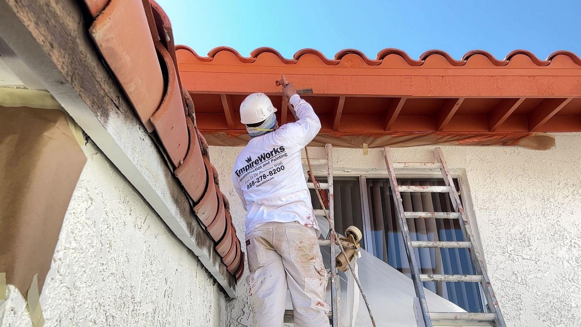 A person in a hard hat and painting clothes on a ladder painting a roof trim.