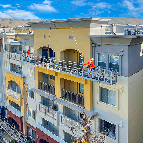 Construction workers on scaffolding, painting a multi-story apartment building exterior.