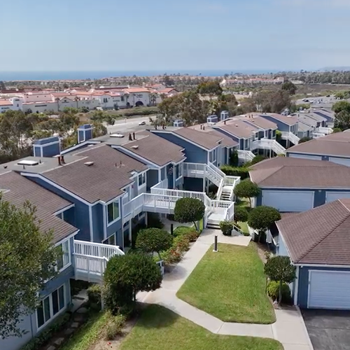 Aerial view of blue apartment complex with brown roofs, white balconies, and green lawns, with ocean view in the background.
