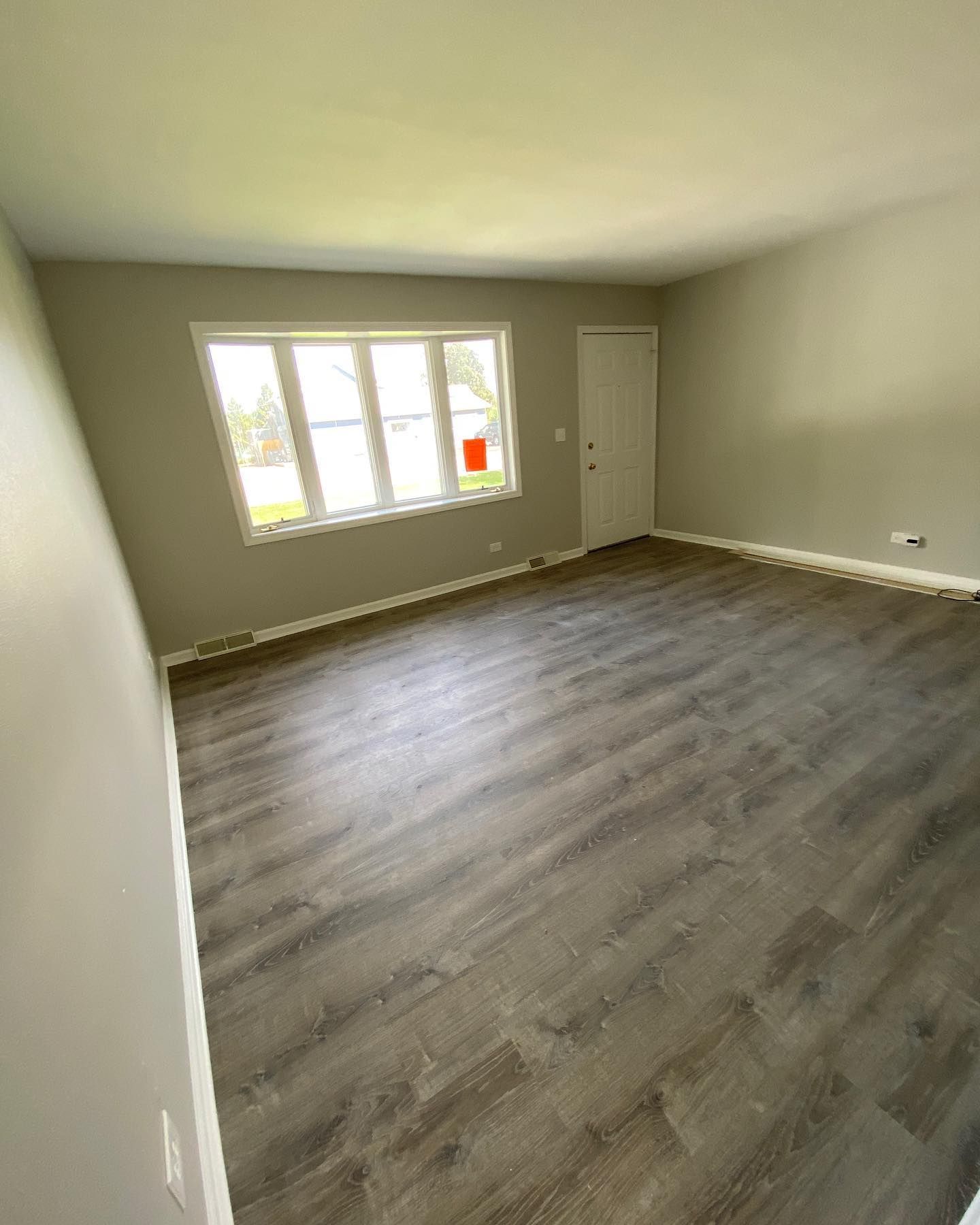 Empty living room with wood-look flooring, light gray walls, large window, and white door.