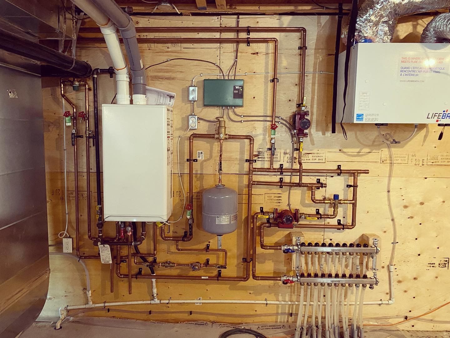 A boiler is sitting on a wooden wall next to a bunch of pipes.