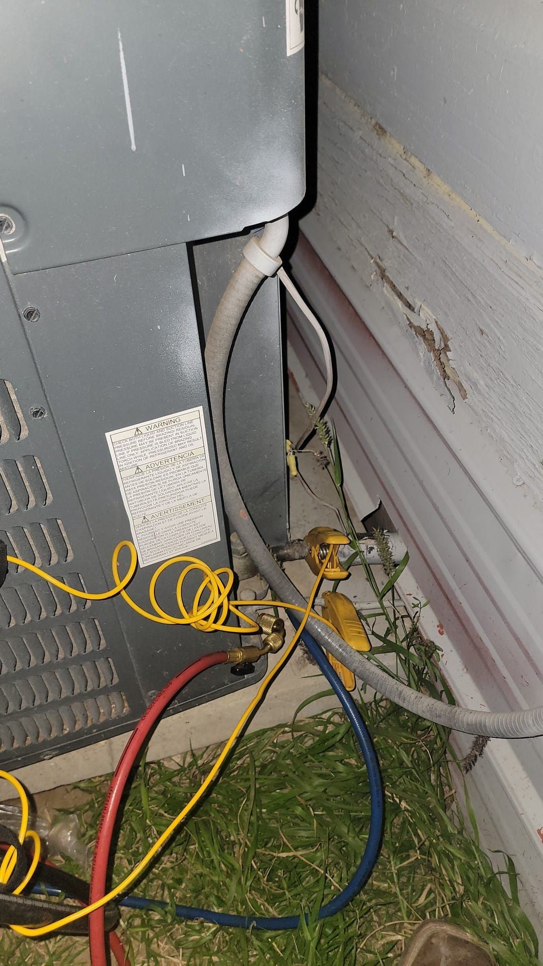 HVAC unit's wires and refrigerant lines near a wall with grass. Yellow, blue, and red tubes.