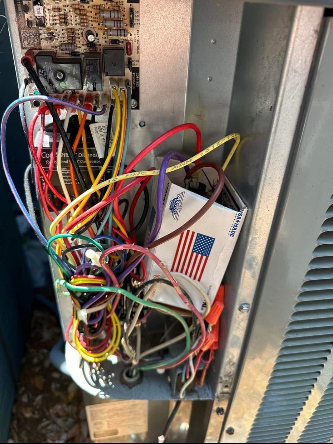 Electrical panel of an HVAC unit, with tangled wires in various colors. A circuit board and capacitor are visible.