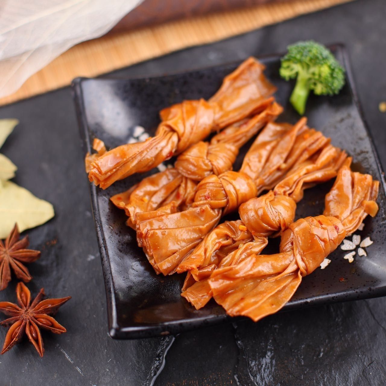 Orange candy wrappers on a black plate with star anise, cinnamon, and a broccoli floret on a dark table