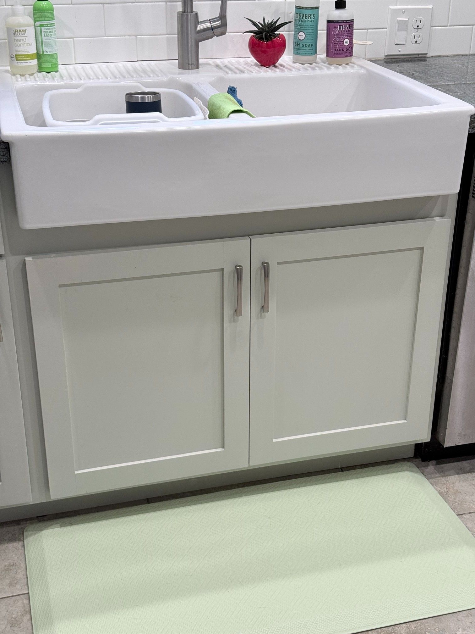 White farmhouse sink above a cabinet, green mat on floor, cleaning supplies and a plant on the counter.