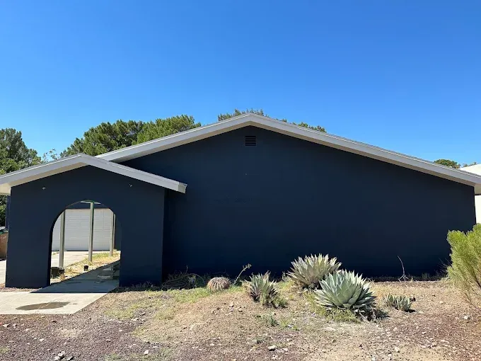 Dark blue house with white trim, entryway arch, and desert landscaping under a blue sky.