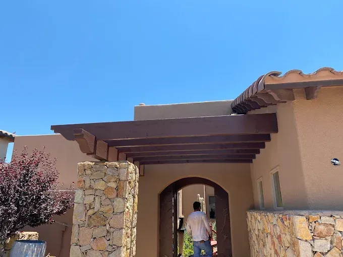 Man walking into an archway of a tan building with a wooden pergola under a blue sky.