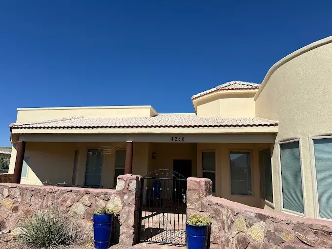 Tan stucco house with rock fence, blue pots, and black gate under a clear blue sky.