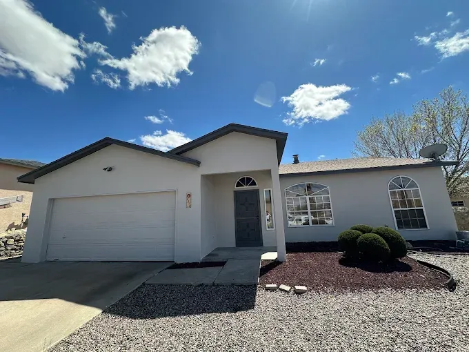 Light gray house with a two-car garage, arched windows, and gravel landscaping under a bright blue sky.