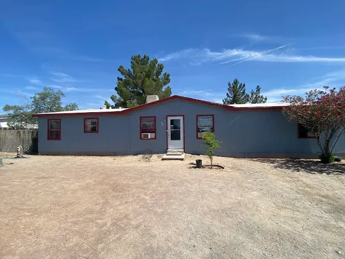 A one-story house with a blue exterior, red trim, and a gravel yard under a blue sky.