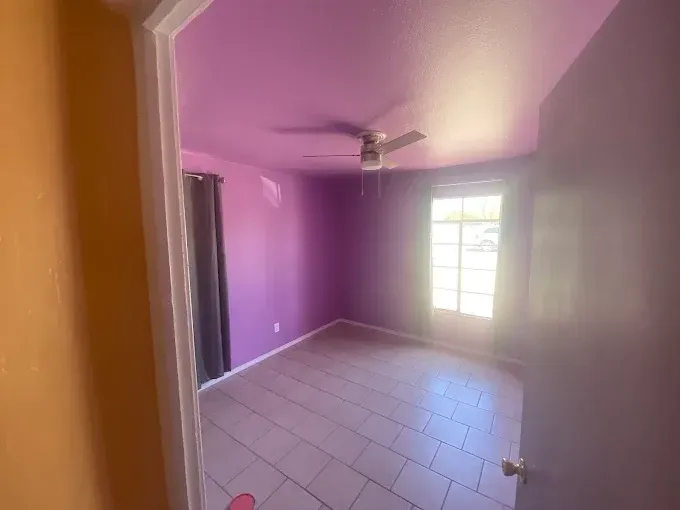 A small purple bedroom with a ceiling fan, a window, and tiled floor, viewed from a doorway.