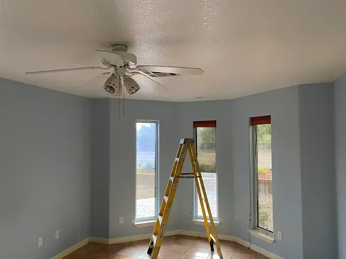 Blue-painted room with a ceiling fan, windows, and a yellow ladder; view of a residential interior.