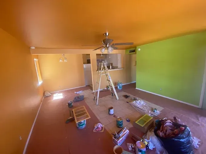 Interior of a room being painted. Orange walls and ceiling, green accent wall, ladder, paint supplies.