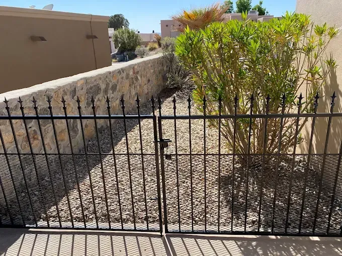 Black wrought-iron gate in front of a gravel yard with a stone wall, plants, and a building in the background.