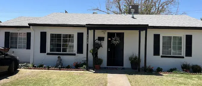 White house with black shutters, a black door, and a gray roof on a sunny day.