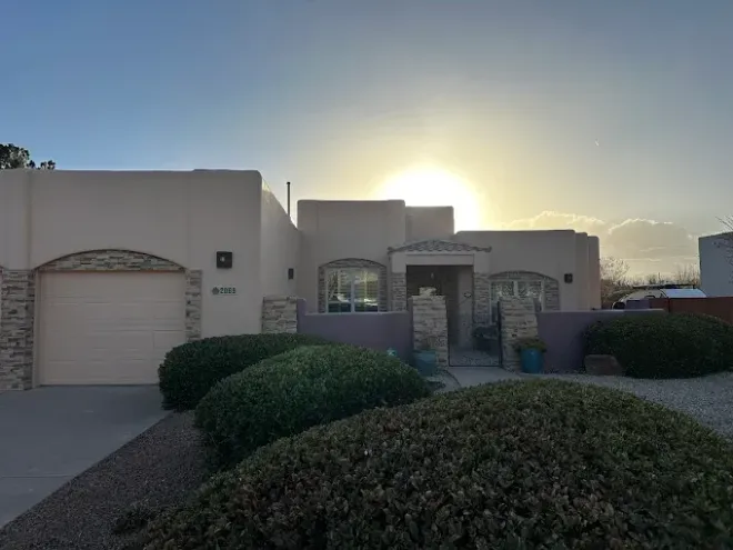 Beige stucco house with stone accents, front yard with bushes, setting sun.