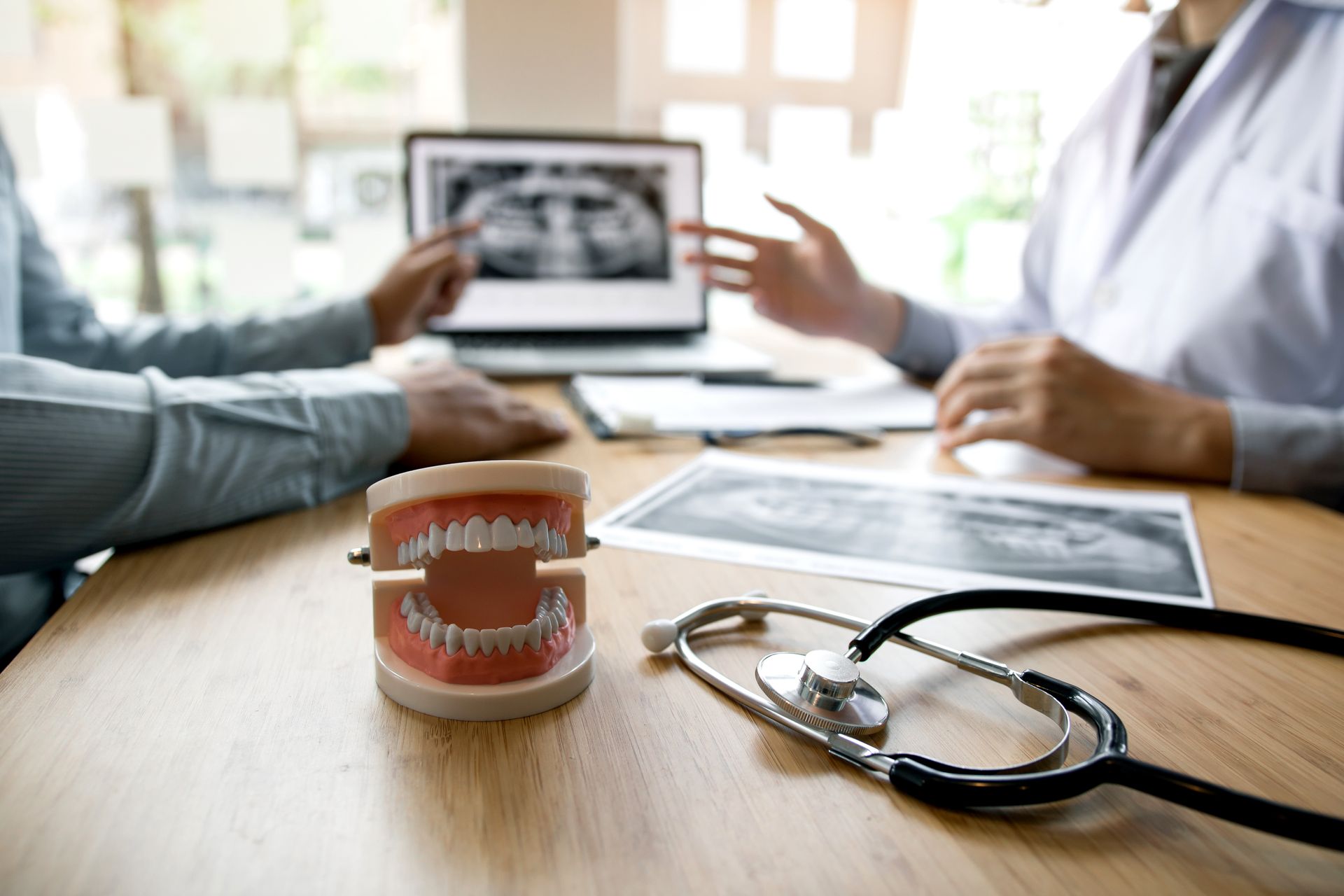 Dentist pointing at X-ray on laptop, consulting patient. Teeth model and stethoscope on table.
