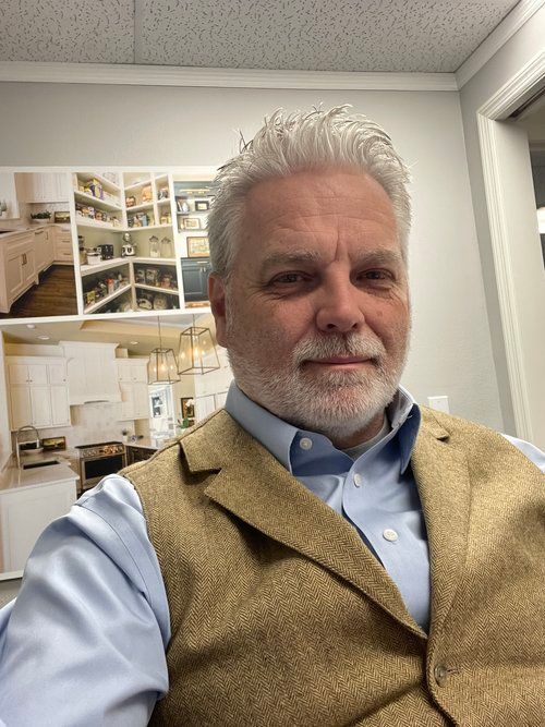A man with a beard is wearing a tan vest and a blue shirt.