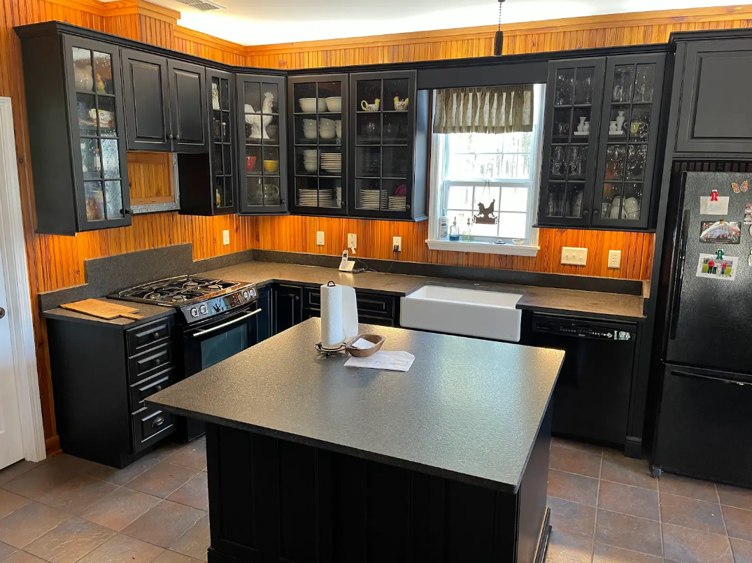 A kitchen with black cabinets , a black refrigerator , a stove , a sink , and a large island.