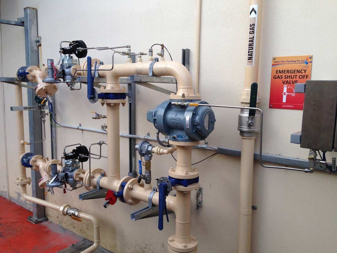 Pipes and valves, beige with blue accents, against a white wall. An emergency shut-off sign is visible.