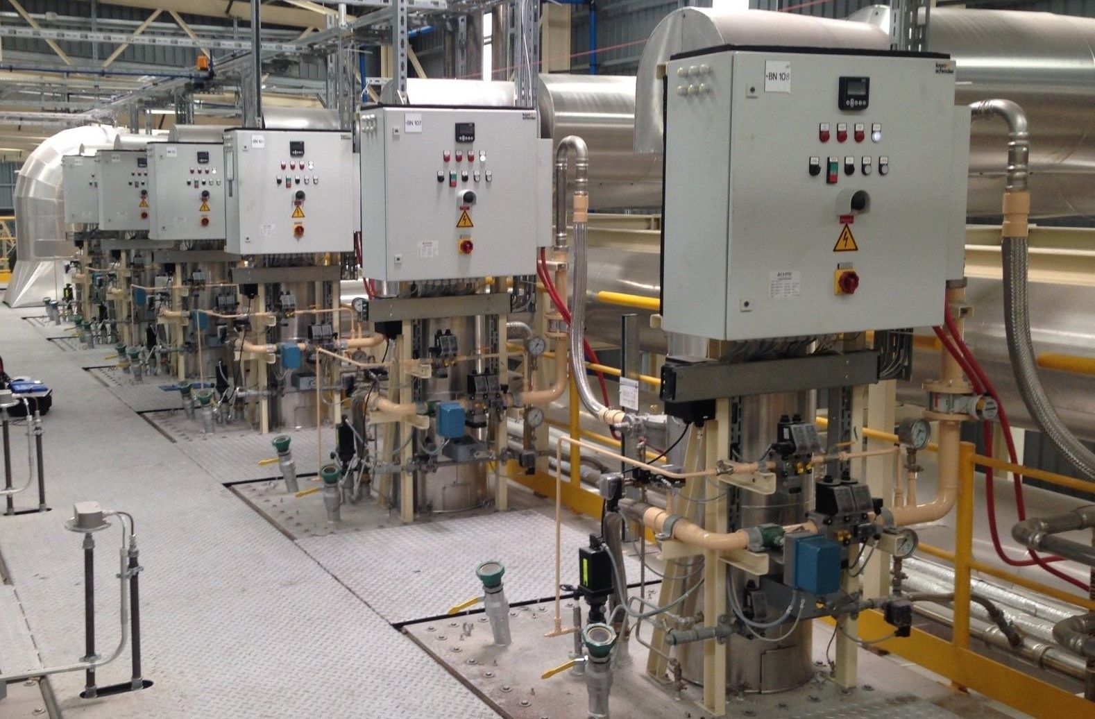Row of industrial control panels and equipment in a factory setting, with piping and machinery.
