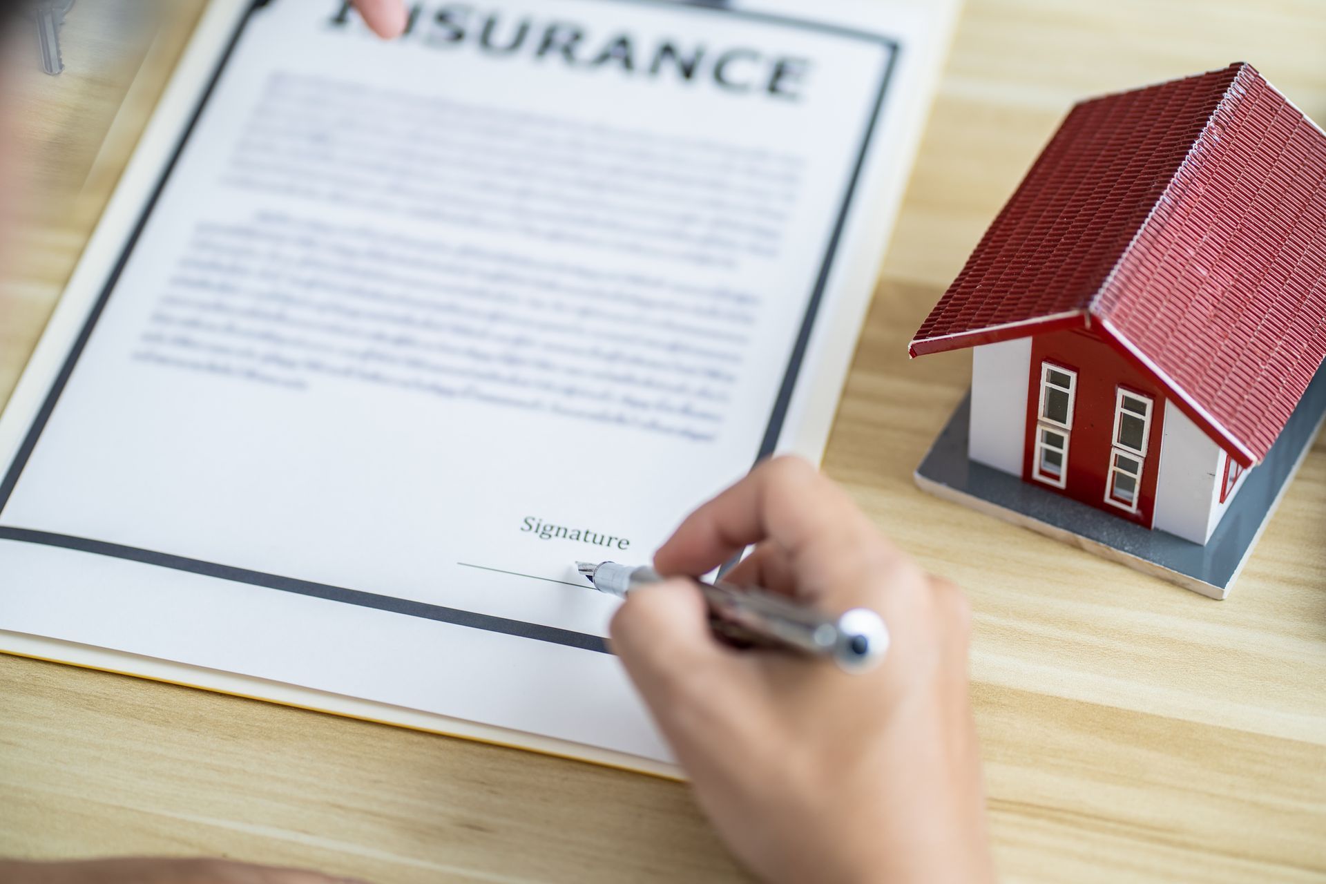 Person signing a home insurance document next to a miniature house.