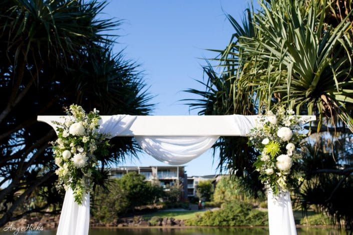 A White Arch With Flowers on It is Surrounded by Trees and a Body of Water — Suncoast Flowers in Birtinya, QLD