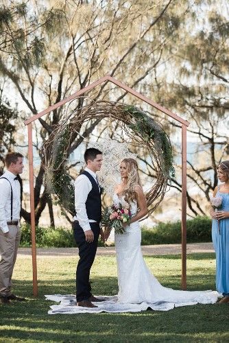 A Bride and Groom Are Standing Under a Copper Arch During Their Wedding ceremony — Suncoast Flowers in Birtinya, QLD