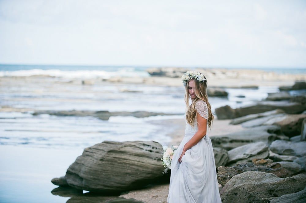 A Woman in a White Dress and Flower Crown is Standing on a Rocky Beach — Suncoast Flowers in Birtinya, QLD