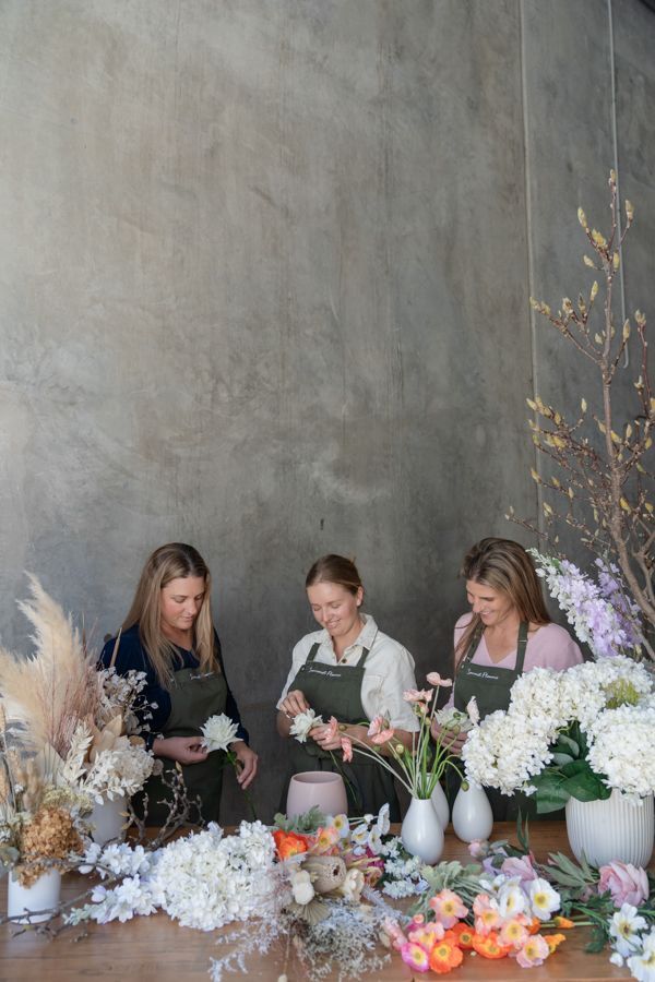 Three Women Are Sitting at a Table With Vases of Flowers — Suncoast Flowers in Birtinya, QLD
