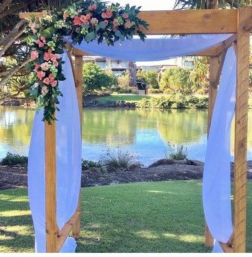 A Wooden Arch Decorated With Flowers and White Curtains is in Front of a Lake — Suncoast Flowers in Birtinya, QLD