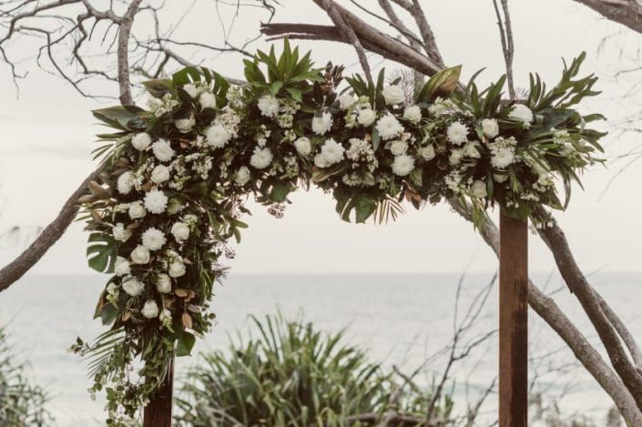 A Wooden Arch Decorated With White Flowers and Leaves on a Beach — Suncoast Flowers in Birtinya, QLD
