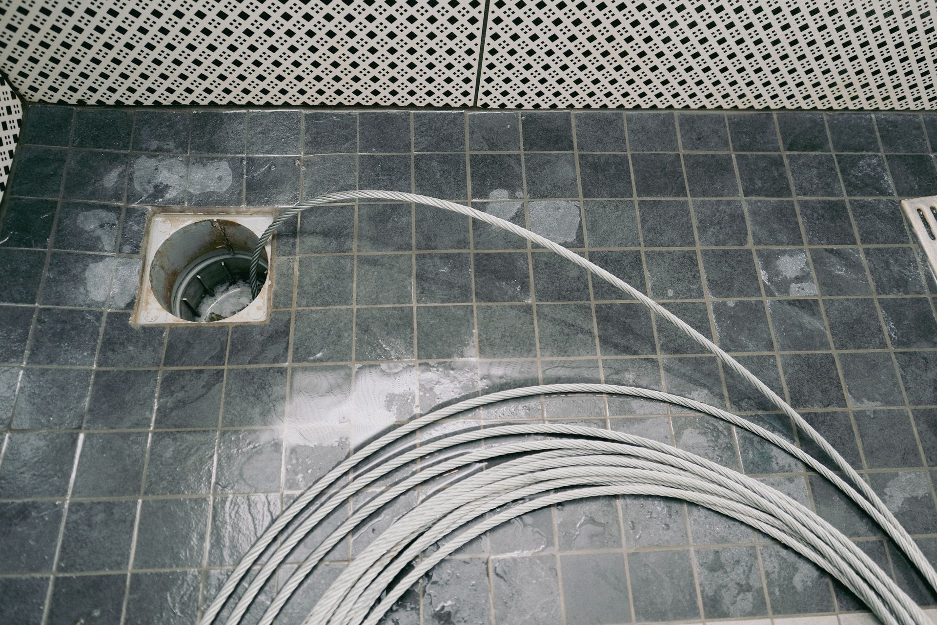 Coiled gray cable on tiled floor, leading to a drain in a bathroom-like setting.