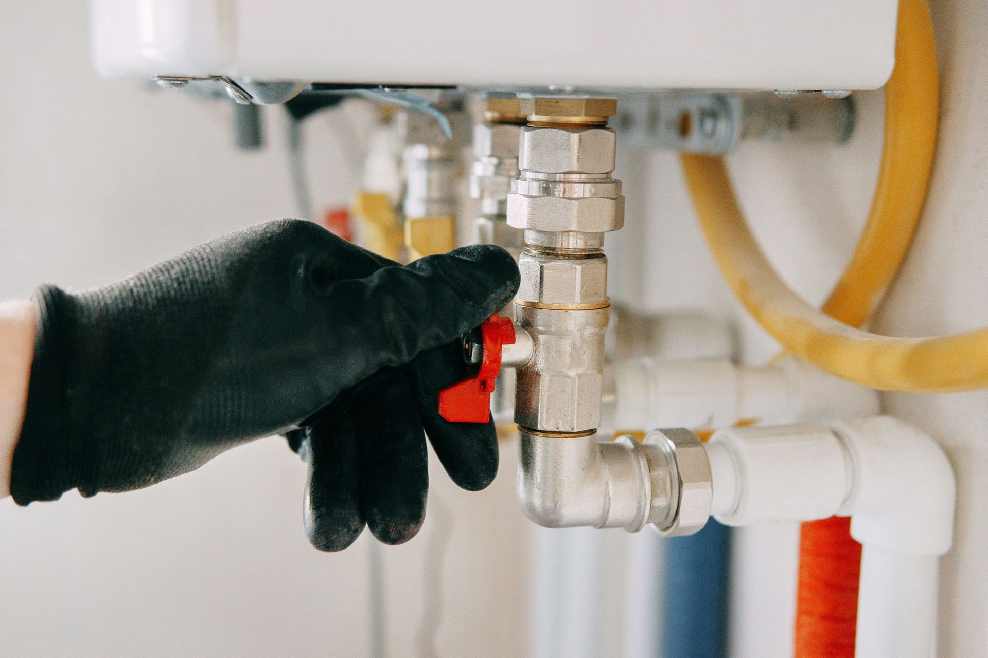 Gloved hand turns a red valve on silver pipes near a water heater, indoors.